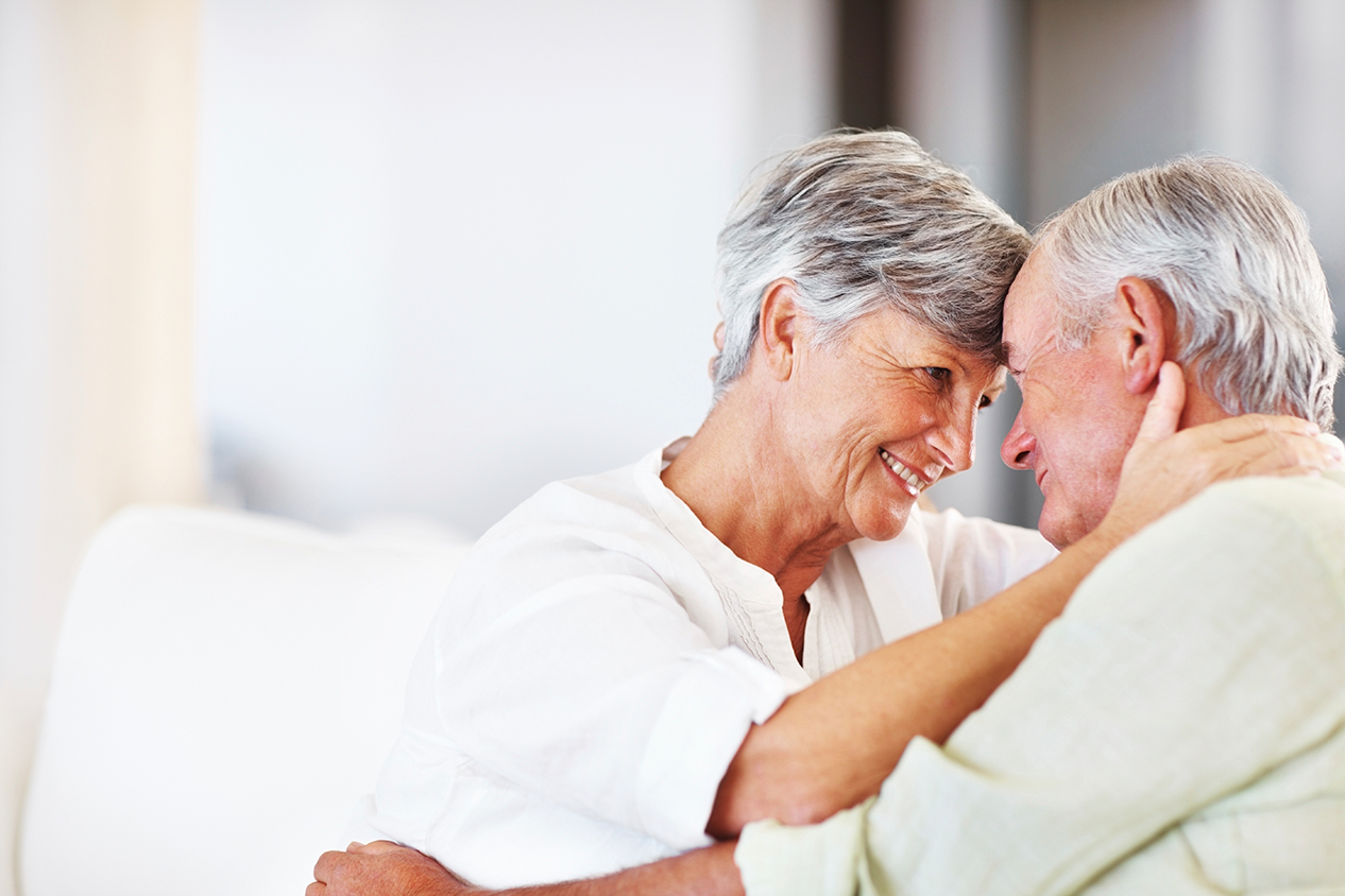 Elderly lady with her hands around elder man's neck smiling 